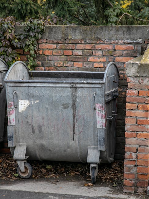 A large, weathered metal waste bin with wheels positioned against a brick wall on a pavement outside a residential property. The bin has visible signs of rust and dirt, with some faded pink and white stickers or markings on its sides. Behind the bin, there is a low brick wall topped with green foliage and bushes, indicating an outdoor setting. This scene illustrates typical waste disposal as part of a home relocation or moving process, where waste materials are temporarily stored before collection. The environment suggests an urban or suburban area with clear space for loading or cleaning during packing and moving activities, often managed by a professional removals service such as Man With a Van Barnsbury.