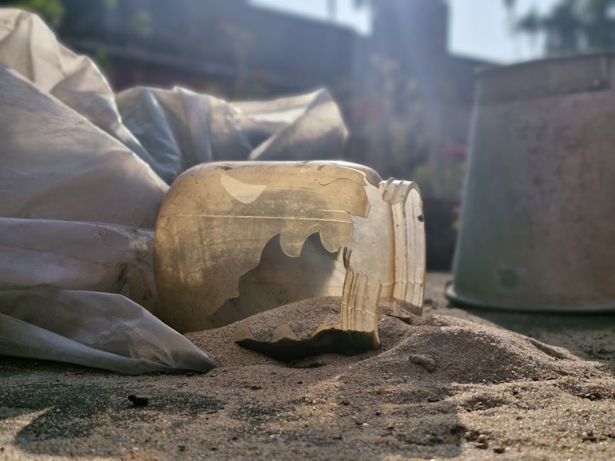 A close-up image shows a broken, partially buried plastic bottle cap lying on sandy ground, with some sand spilled around it. In the background, there are several large, partially covered white and green plastic-wrapped packages, possibly used for packing items during a house removal or relocation. A silver-grey, rounded device or container is visible behind the packages, partially obscured by the sand and plastic coverings. The scene is outdoors, illuminated by natural daylight, with a blurred view of garden elements or surroundings in the distance. This setting illustrates packing or disposal steps during a home relocation process, aligning with services offered by Man With a Van Barnsbury in furniture transport and moving logistics.