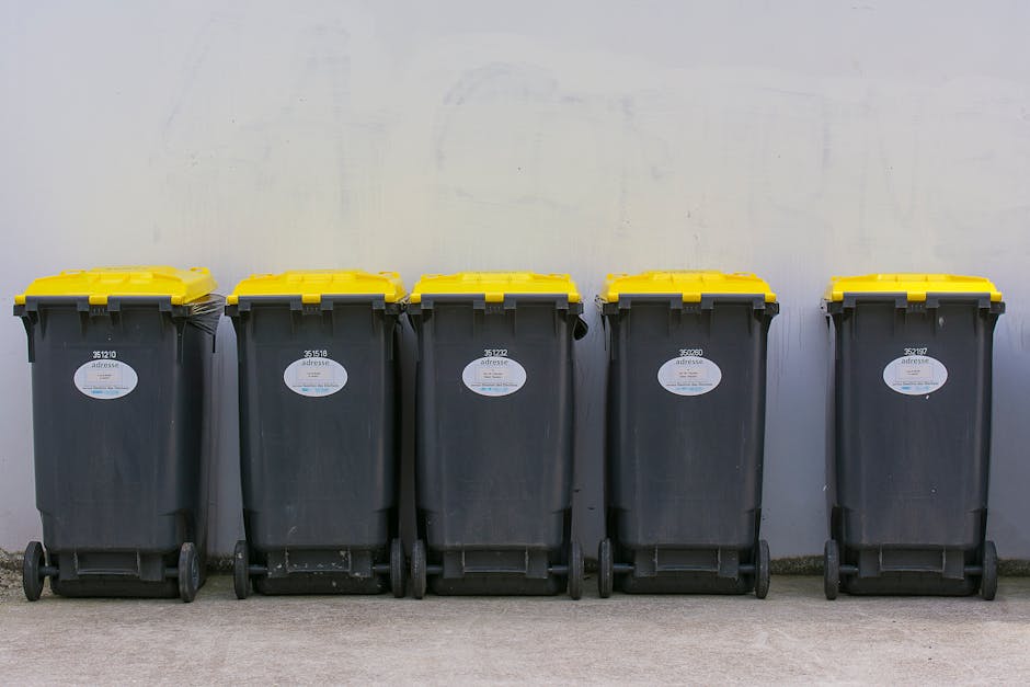 A large, weathered metal waste bin with wheels positioned against a brick wall on a pavement outside a residential property. The bin has visible signs of rust and dirt, with some faded pink and white stickers or markings on its sides. Behind the bin, there is a low brick wall topped with green foliage and bushes, indicating an outdoor setting. This scene illustrates typical waste disposal as part of a home relocation or moving process, where waste materials are temporarily stored before collection. The environment suggests an urban or suburban area with clear space for loading or cleaning during packing and moving activities, often managed by a professional removals service such as Man With a Van Barnsbury.