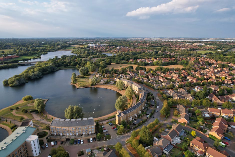 Aerial view of a suburban residential area with a large circular lake surrounded by a curved row of terraced houses, some with chimneys and pitched roofs, and a parking lot with numerous cars adjacent to the buildings. The area features lush green trees and landscaped gardens around the houses, with additional water bodies and greenery visible further in the background. The scene is captured during daylight hours with partly cloudy sky overhead. This setting is typical of a peaceful home relocation environment, where furniture transport and packing essentials are managed around the natural and built environment, consistent with services offered by Man With a Van Barnsbury for house removals and moving logistics.