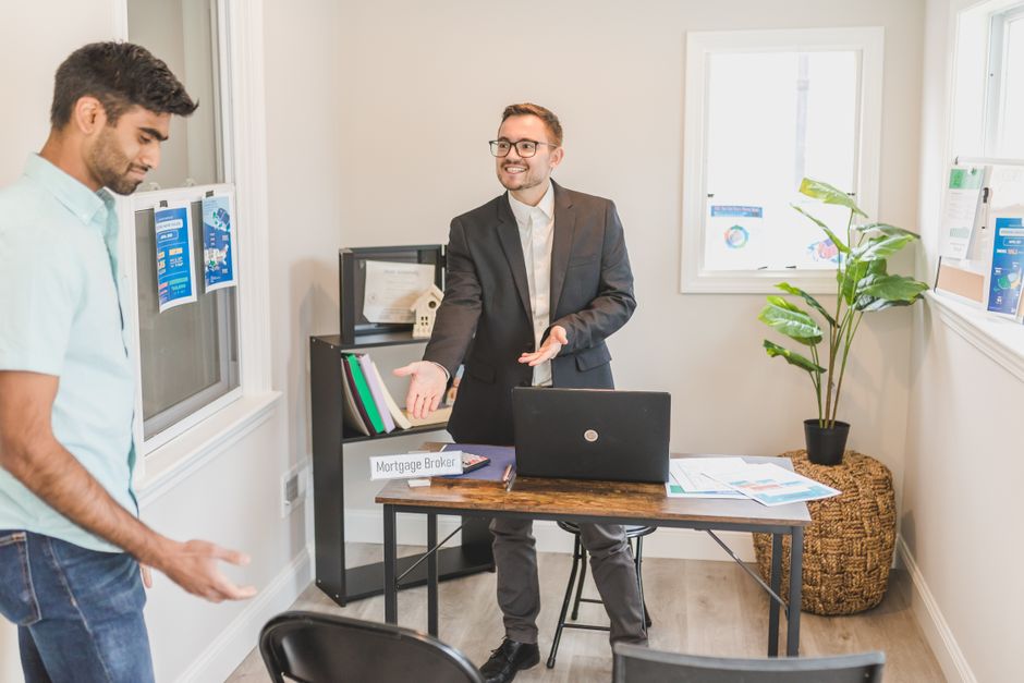 In a small office space with white walls and two windows letting in natural light, a man dressed in a light blue shirt and jeans stands on the left side, looking down at the floor. On the right, a man wearing a dark grey suit, white shirt, and glasses is gesturing with his hands as he speaks, engaging in conversation. The desk in front of him has a closed black laptop and several documents spread out, indicating a discussion about house removals or moving logistics. Behind the suited man, a black shelving unit holds folders, a small model house, and framed certificates, with a small sign labeling him as a 'Mortgage Broker.' A potted plant with large green leaves sits on a woven basket to the side of the window, adding a touch of greenery to the space. The environment suggests a professional meeting related to property transactions or home relocation planning, as part of the broader moving or furniture transport process that Man With a Van Barnsbury might provide.