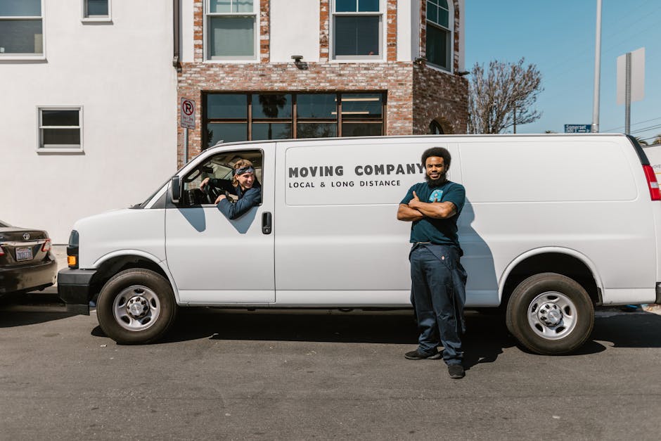 A male furniture removalist with dark curly hair, wearing a navy blue uniform, stands with his arms crossed next to a white moving van branded with 'MOVING COMPANY LOCAL & LONG DISTANCE' on the side, parked on an urban street. The van's sliding door is open, revealing a female removal worker with glasses and a headband inside, who is loading or unloading cardboard boxes and wrapped furniture, including a wooden chair and plastic-wrapped items. The scene takes place near a brick and white-painted residential building with multiple windows, some partially open, and a sign indicating parking restrictions visible above the vehicle. The environment is well-lit with natural daylight, and additional parked cars are visible along the street, with a clear blue sky overhead. The setting reflects a home relocation or furniture transport process typical for professional removals, and the image overall highlights the logistics involved in packing and loading during a house move, as handled by Man With a Van Barnsbury.