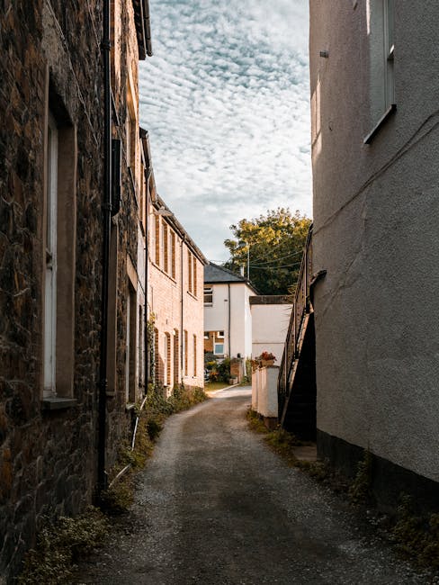 A narrow residential alleyway with a gravel surface, flanked by three-storey stone and brick terraced houses on the left and a white-painted building on the right. The alley leads to a small open area with a glimpse of modern houses in the distance. The scene is captured during daylight with partly cloudy skies overhead. The right side of the image shows a staircase leading up the white building, while the left side features windows and exterior walls of the older houses. This setting illustrates typical urban residential streets suitable for home relocation and furniture transport. Man With a Van Barnsbury, a professional removals company, would utilize such alleyways to plan efficient packing and loading processes during house moves across narrow streets like Barnsbury Road.