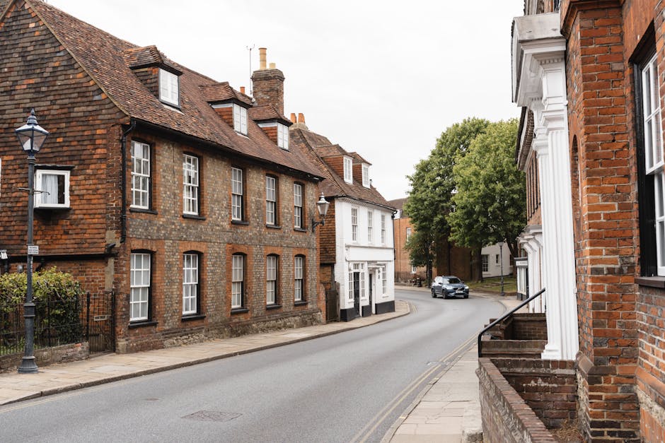A quiet residential street showing traditional brick houses with pitched roofs and multiple chimneys, typical of a historic UK neighbourhood. The pavement runs alongside the buildings, with a vintage-style black lamppost visible near the curb. On the right, part of a brick house with white decorative architectural features around the window and stairs leading up to the entrance can be seen. The street curves gently to the left, and a silver car is driving along the road in the distance. Large green trees provide some natural foliage, shading parts of the scene. This setting illustrates a typical environment where home relocation services by Man With a Van Barnsbury are often required, highlighting the narrow street layout and urban residential architecture suitable for discreet and efficient furniture transport, packing, and loading processes involved in house removals.