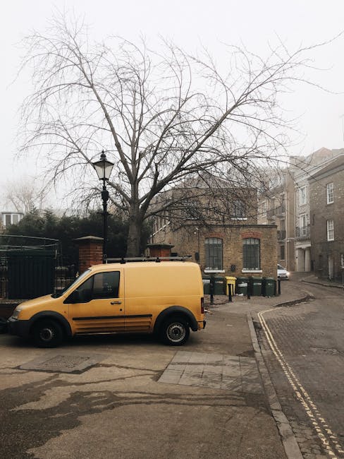 A yellow delivery van is parked on a narrow urban street with double yellow lines, positioned close to the pavement curb. Behind the van, there is a leafless tree with numerous thin branches, and it is set against a cloudy, overcast sky. To the right of the van, a building with a brick facade and dark window frames is visible, along with a black metal fence and green wheelie bins lined up along the edge of a property. Further down the street, residential buildings with beige and brick exteriors are seen, with some windows reflecting local street lighting. The street appears to be part of a residential area in Barnsbury, with a slight incline leading to more buildings in the distance. The scene is muted due to the overcast weather, and the overall environment suggests it may be part of a home relocation or furniture transport process, as operated by Man With a Van Barnsbury, highlighting the typical loading area for house removals and moving services.