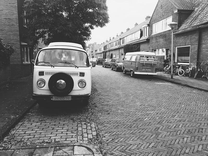 A yellow delivery van is parked on a narrow urban street with double yellow lines, positioned close to the pavement curb. Behind the van, there is a leafless tree with numerous thin branches, and it is set against a cloudy, overcast sky. To the right of the van, a building with a brick facade and dark window frames is visible, along with a black metal fence and green wheelie bins lined up along the edge of a property. Further down the street, residential buildings with beige and brick exteriors are seen, with some windows reflecting local street lighting. The street appears to be part of a residential area in Barnsbury, with a slight incline leading to more buildings in the distance. The scene is muted due to the overcast weather, and the overall environment suggests it may be part of a home relocation or furniture transport process, as operated by Man With a Van Barnsbury, highlighting the typical loading area for house removals and moving services.