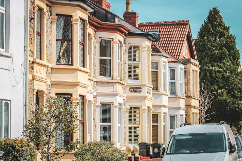Photograph of a residential street showing a row of Victorian-style terraced houses with bay windows, decorative brickwork, and red-tiled roofs. The houses are illuminated by daylight, and trees and greenery are visible in the background. In the foreground, part of a white van with a roof rack is parked on the pavement, indicating the start of a home relocation process. Visible items include cardboard boxes, wrapped furniture, and packing materials placed near the entrance of one house, suggesting ongoing packing or loading activities. A small trolley or dolly might be seen in use, with some furniture or boxes being moved towards the van, which is positioned close to the property for loading. The setting highlights typical elements involved in furniture transport and moving logistics, relevant to services like those offered by [COMPANY_NAME] for local removals in N1 Barnsbury, including packing, lifting, and transport of household items.