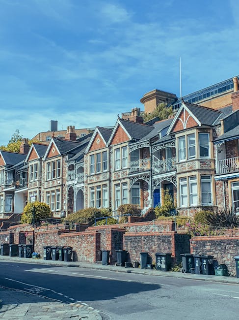 A row of Victorian terraced houses with decorative brickwork, bay windows, and intricate ironwork balconies, situated on a residential street in Barnsbury. The houses are elevated on brick retaining walls with small gardens and shrubs in front of each property. A parked van is visible alongside the curb, and the sky above is clear with a few scattered clouds, indicating a bright, sunny day. This scene exemplifies typical house exteriors in Barnsbury, relevant to home relocation and furniture transport services provided by Man With a Van Barnsbury, situated in the context of house removals and moving logistics.