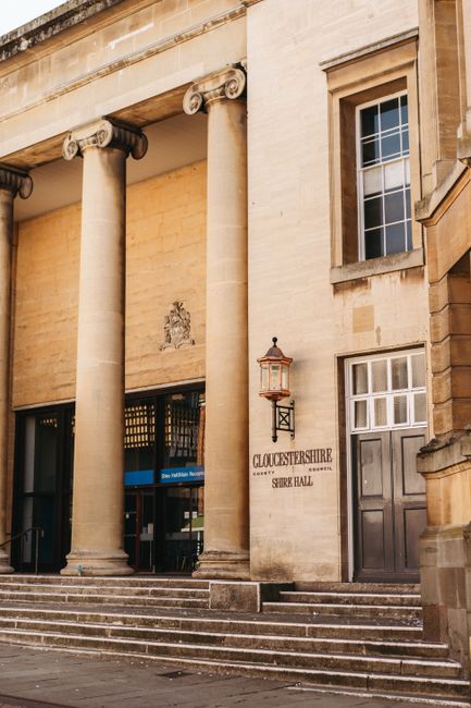 The entrance of a historic stone building featuring classical architectural elements, including two large, fluted columns with ornate capitals supporting a prominent entablature. The facade is made of light-colored stone with decorative shields and carvings above the entrance. To the right of the columns, there is a dark wooden door with a brass handle, and above it, a window with multiple small panes. A traditional lantern-style outdoor light fixture is mounted on the wall near the door, illuminating the building's signage which reads 'GLOUCESTERSHIRE SHIRE HALL.' The steps leading up to the entrance are made of stone, providing access for visitors. The scene is well-lit, highlighting the building’s historic architectural details, suitable for a context involving heritage buildings or official municipal services related to house removals and property relocation, such as those managed by Man With a Van Barnsbury.