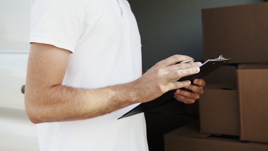 Close-up of a person wearing a white t-shirt holding a clipboard and pen, preparing to record information during a home relocation or furniture transport process within a residential property. In the background, there are large cardboard moving boxes stacked on top of each other, some wrapped with plastic or packing materials, indicating packing and packing materials used for house removals. The scene appears to take place inside a house, near a doorway or hallway, with ambient indoor lighting. The individual is likely part of a professional moving service such as Man With a Van Barnsbury, involved in organising or checking items for a house removal or home relocation. The focus is on the person's hands, clipboard, and the boxed packaging materials, illustrating the packing, loading, and logistics phases typical of furniture transport or house removals.