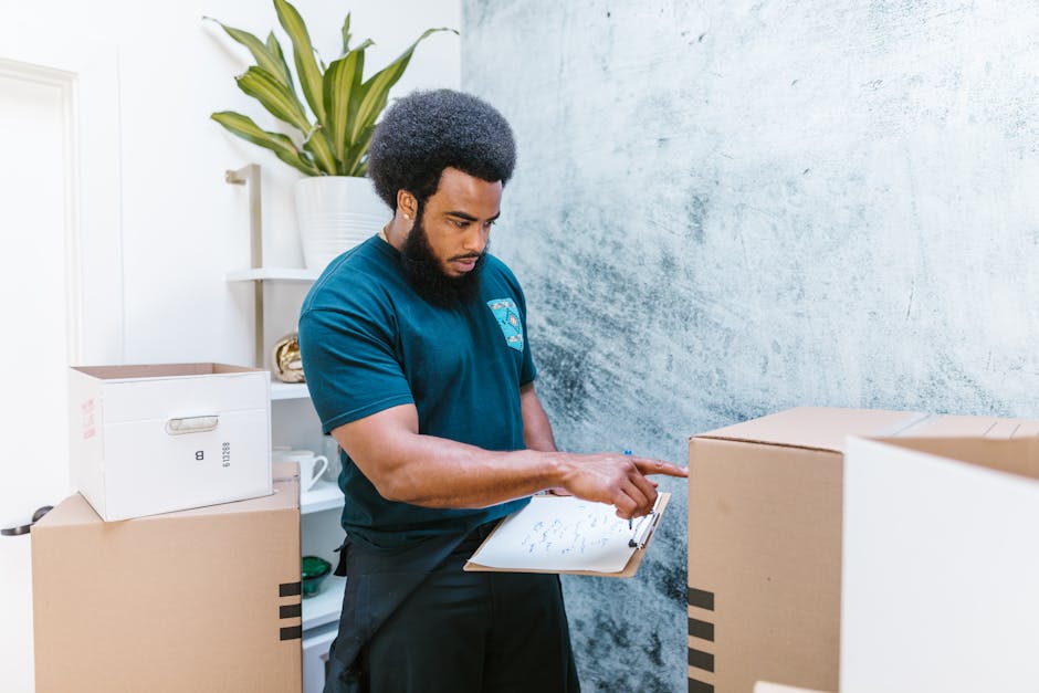 A man with dark skin, curly black hair, and a beard is positioned indoors, surrounded by several cardboard boxes, some unopened and others partially packed. He is holding a clipboard with notes and pointing at a box with his index finger, indicating an organized packing or inventory process. The room features a textured blue wall and a white shelf in the background that holds a large potted plant with long, green leaves. The lighting is bright and natural, illuminating the area clearly. The scene captures the preparation phase of a home relocation, involving careful packing and equipment management. The presence of boxes, packing materials, and the man's focused activity all reflect a typical furniture transport or household moving process, aligning with professional removals services like those offered by Man With a Van Barnsbury, supporting timely and organized moving logistics for clients.