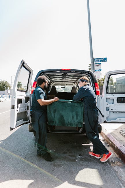 Two movers from Man With a Van Barnsbury are engaged in the loading process of a commercial cargo van, positioned outdoors on a street in Barnsbury. The scene shows them carefully lifting or adjusting a large, dark green piece of furniture, possibly a sofa or a bulky household item, which is partially wrapped in protective fabric or padding to prevent damage during transit. The rear doors of the van are wide open, revealing an interior space designed for moving furniture and boxes, with some additional packaging materials visible inside. The environment is bright and sunny, with a pedestrian lamp post, road sign, and nearby parked vehicles indicating a typical urban setting. The movers are dressed in casual work attire, including safety glasses, and appear focused on ensuring the secure handling of the item for home relocation or furniture transport. This scene exemplifies professional packing and loading procedures used by [COMPANY_NAME] in house removals and moving services in Barnsbury, highlighting their commitment to safe and efficient furniture transport.