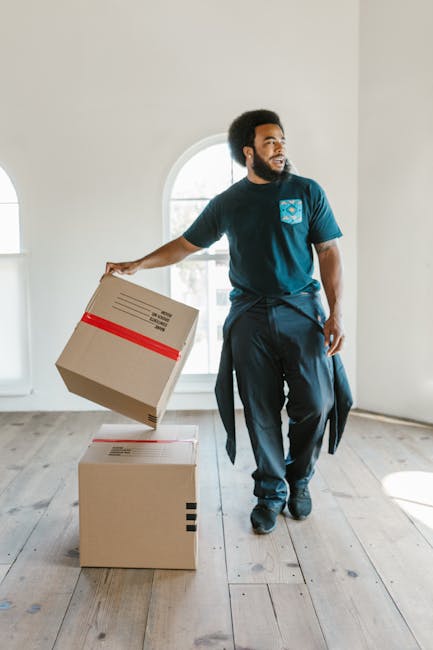 Two movers, dressed in casual dark clothing, are engaged in a home relocation task inside a bright room with wooden flooring and large arched windows. Each mover is positioned on either side of a teal velvet sofa, carefully lifting it from the floor. The man on the left has long hair secured with a headband and is wearing orange and white sneakers, while the man on the right has an afro hairstyle, a beard, and is wearing black shoes. The sofa appears to be a mid-century style with wooden legs and tufted upholstery. Natural light floods the room through the large windows, illuminating the scene and highlighting the careful handling of the furniture. This image illustrates the logistics involved in packing and moving large, bulky furniture as part of a home removal service, with the movers utilizing proper lifting techniques and coordinating their efforts, consistent with professional furniture transport practices. Man With a Van Barnsbury is involved in such removals, ensuring safe and efficient furniture transport during house relocations.