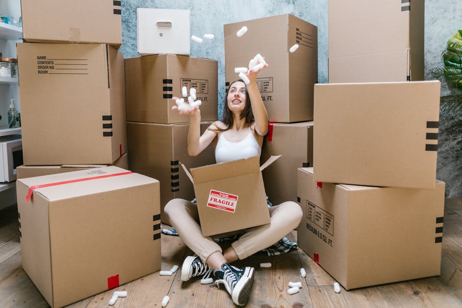 A young woman with long dark hair, wearing a white tank top, beige trousers, and sneakers, is sitting cross-legged on a wooden floor inside a room during a home relocation process. She is surrounded by numerous brown cardboard moving boxes of various sizes, some closed with red tape and others open, revealing packing materials such as foam packing peanuts and bubble wrap. The woman is actively throwing foam packing peanuts into the air, smiling and looking upwards. Behind her, there are more boxes stacked against a textured wall painted in a muted blue tone. To her right, a small section of a white side table and a green potted plant are visible. The scene captures the packing and loading phase of a furniture transport or house move, with [COMPANY_NAME] facilitating the process of packing or organizing items for transportation, likely in coordination with a professional removals service like Man With a Van Barnsbury.