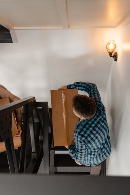 A man wearing a blue and black checked shirt is carrying a medium-sized cardboard box during the home relocation process inside a narrow staircase. The staircase has white walls and a black metal railing, with several stacked cardboard boxes and packing materials visible along the side. The lighting from a wall-mounted sconce illuminates the area. The man is carefully lifting the box, preparing to transport it to a different level. This scene depicts the loading and moving process typical of furniture transport and packing services provided by [COMPANY_NAME], in line with house removals and tight-stair logistics in Barnsbury. The setting reflects an interior space with a modest, clean design, emphasizing the careful handling of household items during professional removals conducted by [COMPANY_NAME].