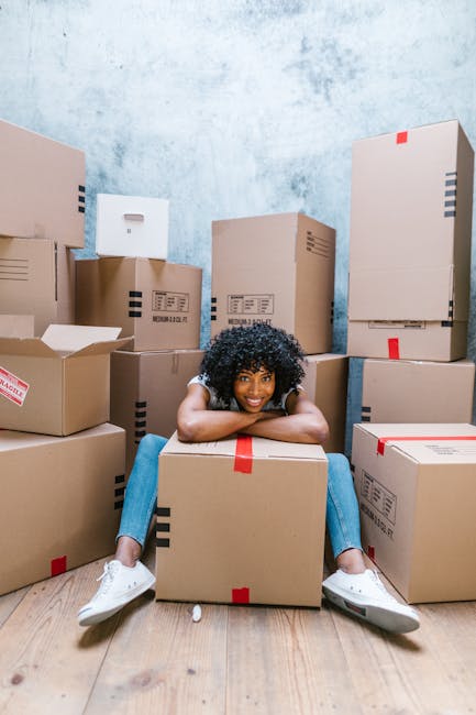 A young woman with long dark hair, wearing a white tank top, beige trousers, and sneakers, is sitting cross-legged on a wooden floor inside a room during a home relocation process. She is surrounded by numerous brown cardboard moving boxes of various sizes, some closed with red tape and others open, revealing packing materials such as foam packing peanuts and bubble wrap. The woman is actively throwing foam packing peanuts into the air, smiling and looking upwards. Behind her, there are more boxes stacked against a textured wall painted in a muted blue tone. To her right, a small section of a white side table and a green potted plant are visible. The scene captures the packing and loading phase of a furniture transport or house move, with [COMPANY_NAME] facilitating the process of packing or organizing items for transportation, likely in coordination with a professional removals service like Man With a Van Barnsbury.