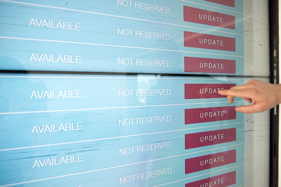 A person pointing at a digital display screen showing multiple rows labeled 'AVAILABLE' and 'NOT RESERVED' in white text, with red buttons labeled 'UPDATE' on the right side of each row. The screen appears to be located in a building lobby or reception area, with reflections of the environment visible on its surface. The background suggests an indoor setting with natural light. This image depicts the process of checking or updating reservation or availability status, relevant to logistics and planning in house removals or moving services, as provided by companies like Man With a Van Barnsbury, supporting efficient home relocation and furniture transport logistics.