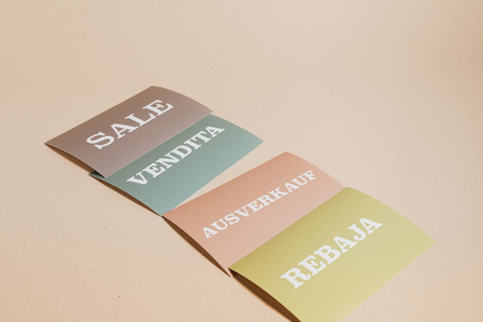 Four rectangular paper signs are laid out in a staggered arrangement on a light beige surface, each featuring bold white text in uppercase. From top to bottom, the signs read 'SALE,' 'VENDITA,' 'AUSVERKAUF,' and 'REBajas,' indicating different languages for sales signage. The signs are in muted pastel colors: gray, green, peach, and yellow, respectively. They are slightly curled at the edges, suggesting they are made of flexible paper or cardboard. The background surface appears smooth and clean, with no other objects visible. This image may be used to illustrate packaging, signage, or promotional materials related to house removals, packing, or moving services—such as those offered by Man With a Van Barnsbury—highlighting the importance of clear communication during a home relocation, including labeling and signage for packing and transport processes.